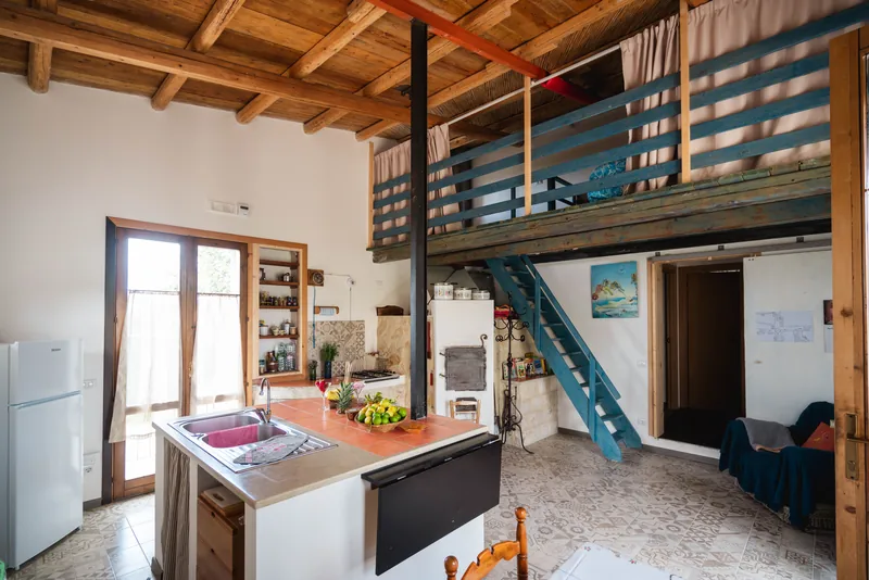Cucina luminosa con isola, travi di legno e scala turchese verso il soppalco